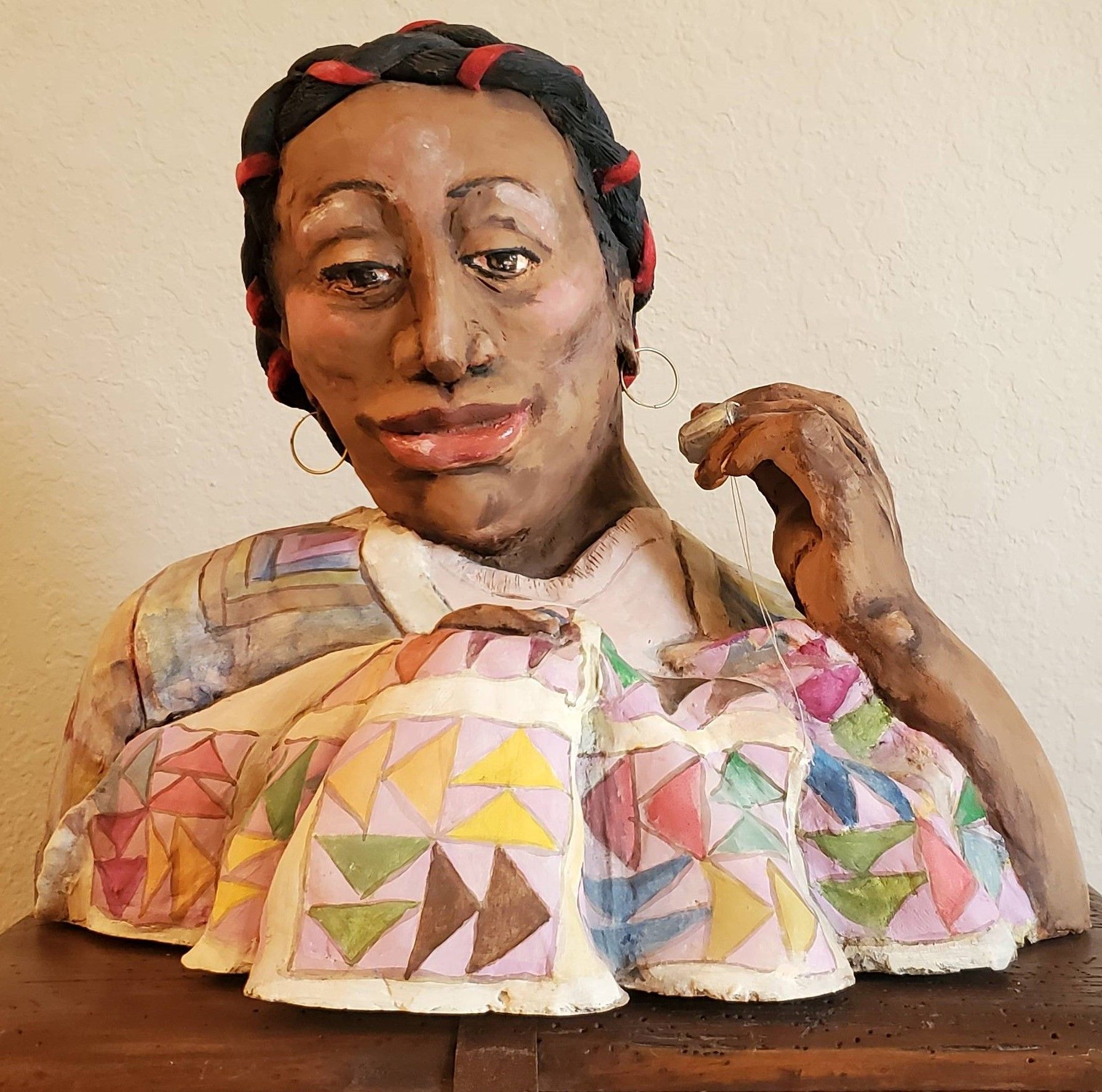 A sculpture of a colorfully dressed woman wearing a thimble and holding a piece of thread while she sews a blanket